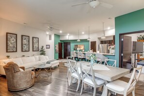 A modern living room with a white sofa, a dining table with chairs, and a kitchen area with a fridge and cabinets.