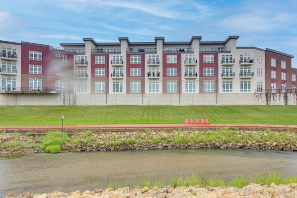 A multi-story residential building with a stone facade, a grassy area in front, and a small body of water. - Rooftop Deck, Great Views! Modern Condo Near Dtwn (Dyersville)