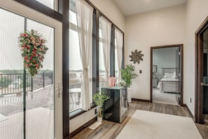 A hotel room with a balcony, a large window with curtains, a small table with a vase, and a bed with a headboard.