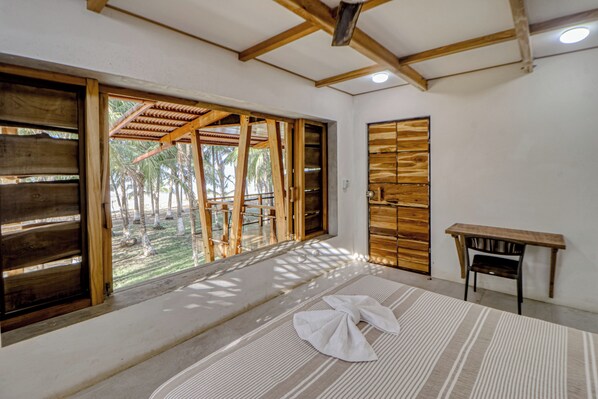 A room with a large window, wooden beams, a bed with a striped bedspread, and a small table.