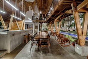 A spacious indoor area with a long wooden table and chairs, a concrete wall, and a view of palm trees outside.