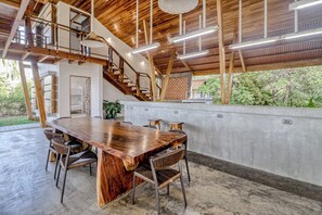 A spacious room with a large wooden dining table, multiple chairs, a staircase, and a view of greenery outside.