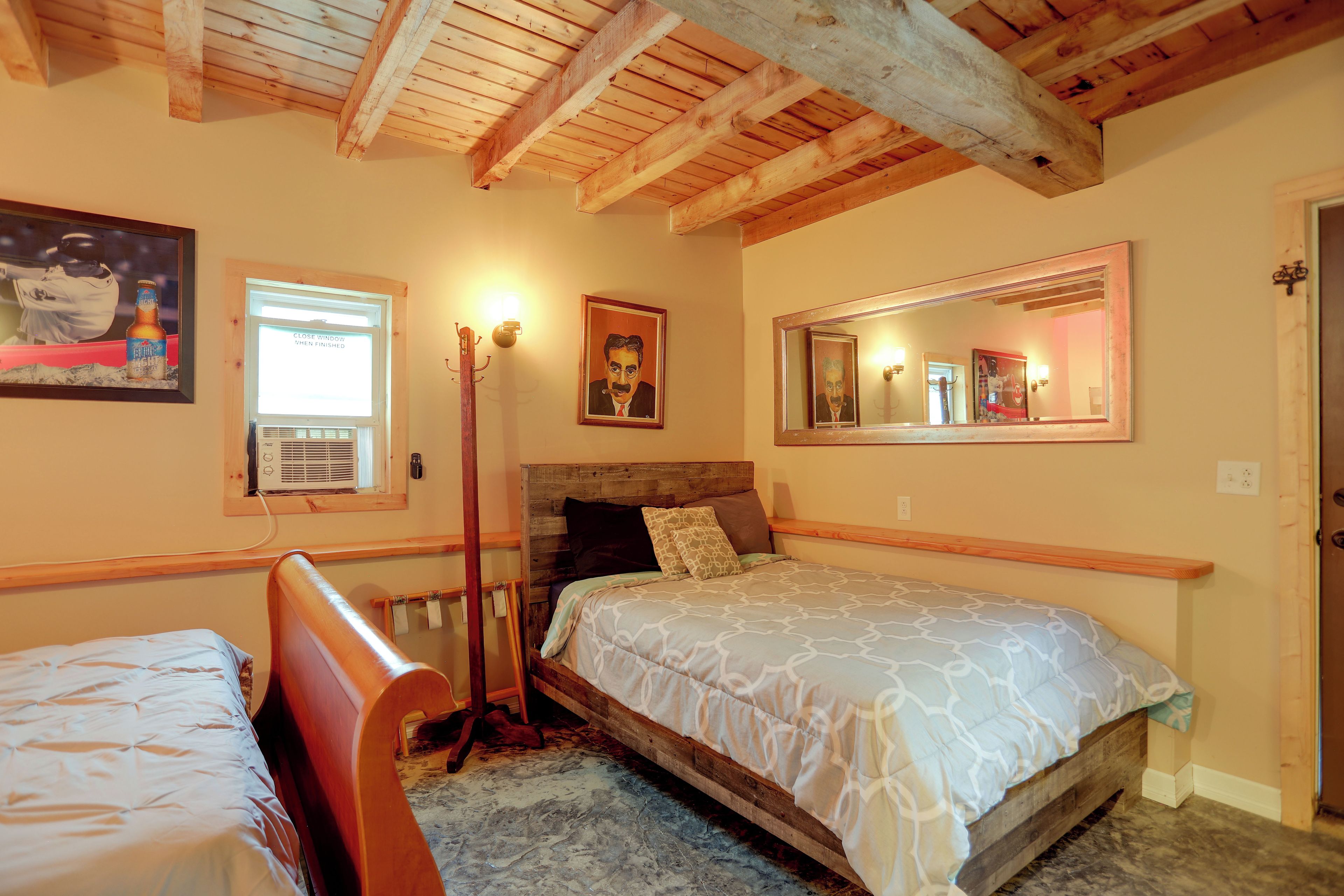 A bedroom with a wooden ceiling, a bed with a patterned bedspread, a bedside table, a chair, a window with a closed blind, and a wall-mounted mirror.