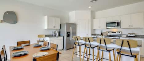 Una cocina moderna con armarios blancos, electrodomésticos de acero inoxidable y una zona de comedor con sillas de madera y platos.