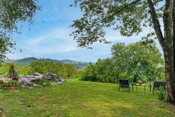 Un espacio al aire libre con césped, dos sillas y una mesa, rodeado de árboles y vistas a colinas.