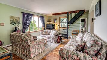 A living room with floral-patterned sofas, a wooden floor, a staircase, and a large window with curtains.