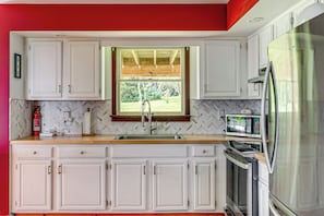 Una cocina con armarios blancos, un refrigerador de acero inoxidable, un microondas y un ventanal con vista a zonas verdes.