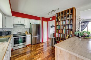 Una cocina con armarios blancos, electrodomésticos de acero inoxidable, piso de madera y una estantería llena de libros.