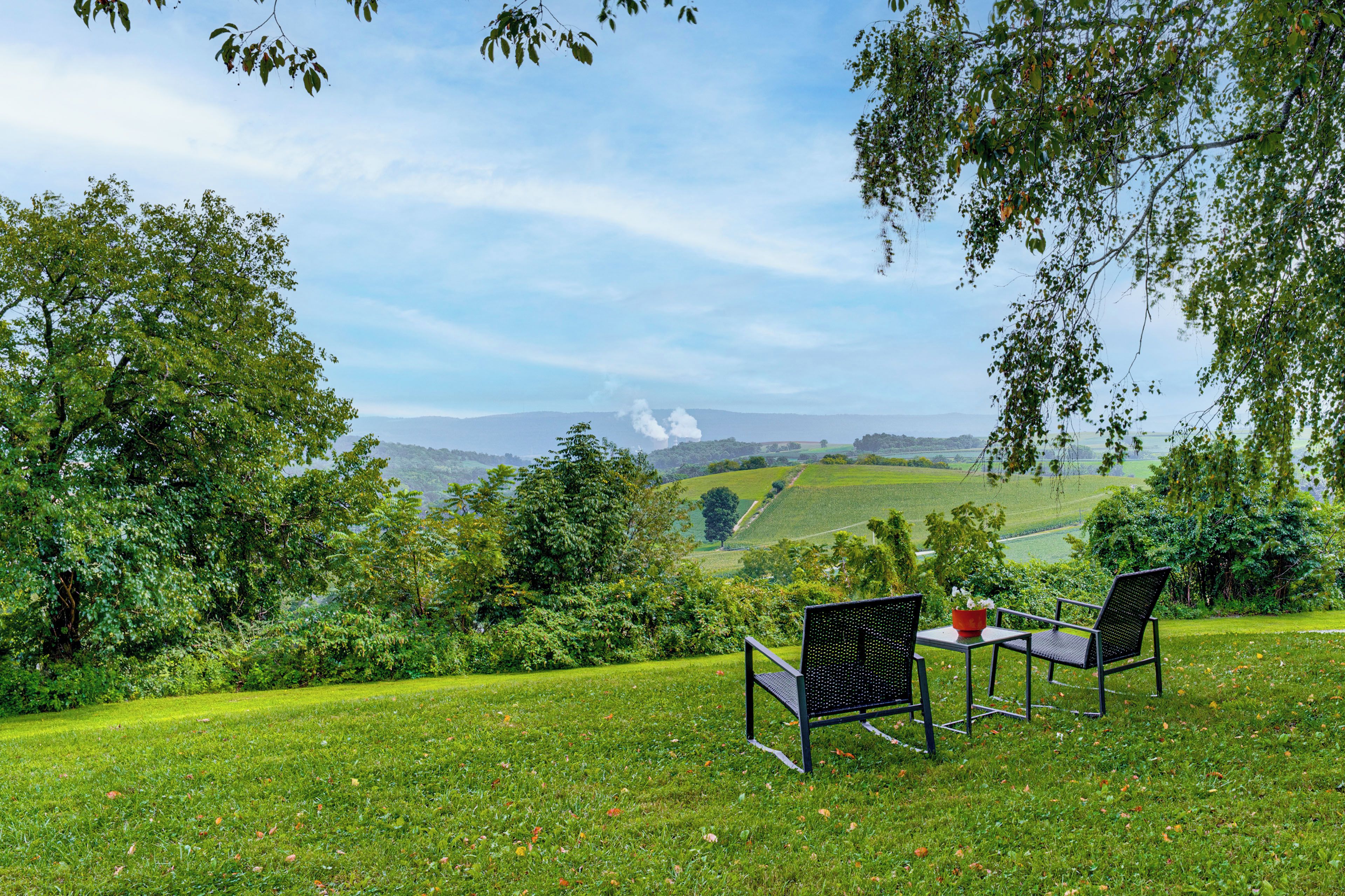 A grassy outdoor area with two chairs and a table, overlooking a scenic landscape with hills and a distant smoke plume.