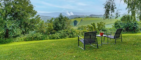 A grassy outdoor area with two chairs and a table, overlooking a scenic landscape with hills and a distant smoke plume.