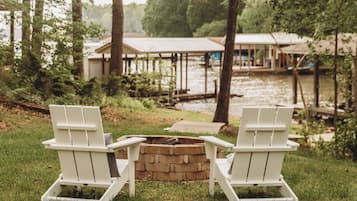 Two white Adirondack chairs facing a brick fire pit with a view of a wooden dock and a building across the water.