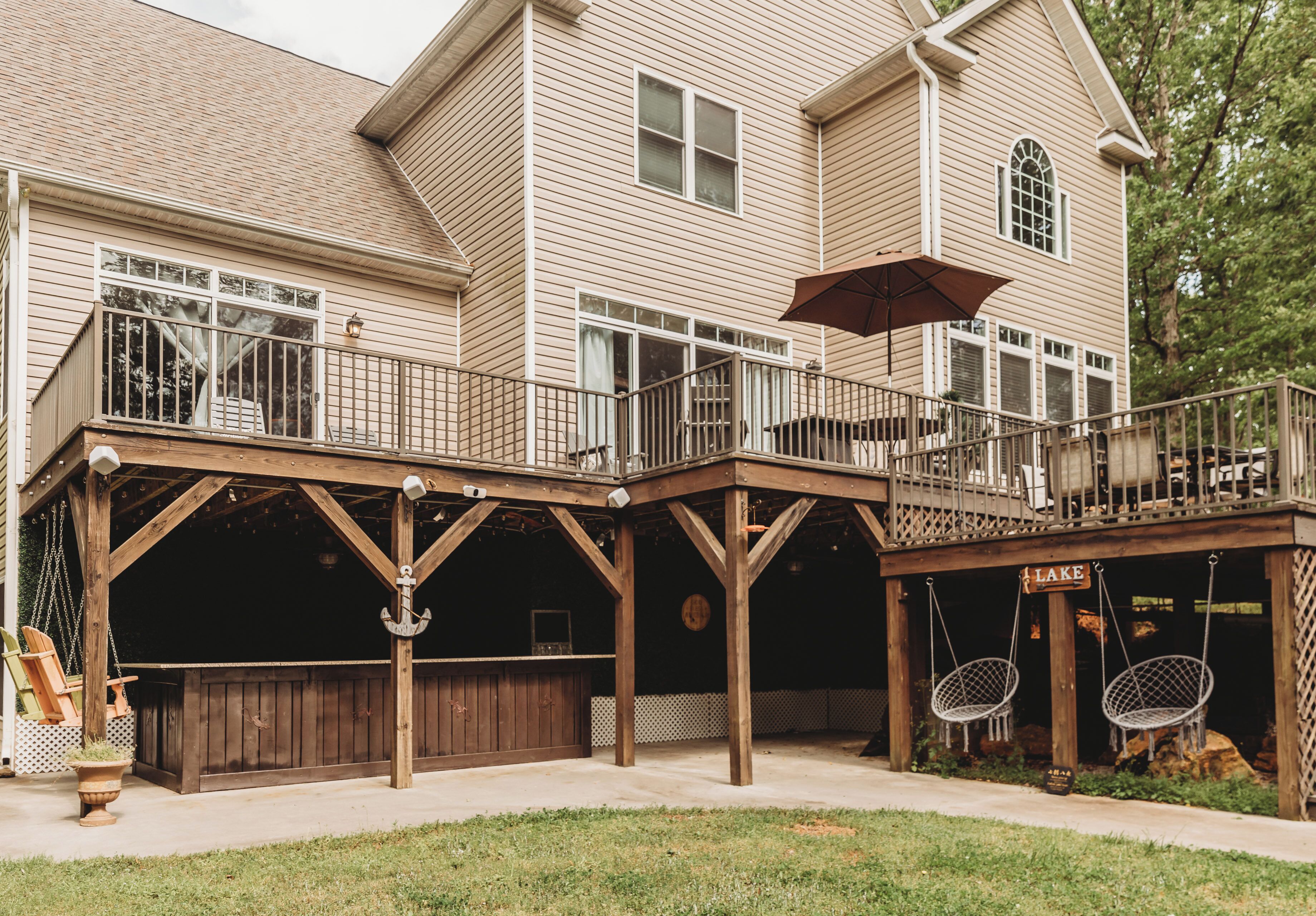 A two-story house with a covered patio area featuring a swing set, a wooden deck, and a sign that reads 'LAKE'.
