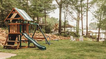 A playground with a slide and swings, surrounded by trees and a building in the background.