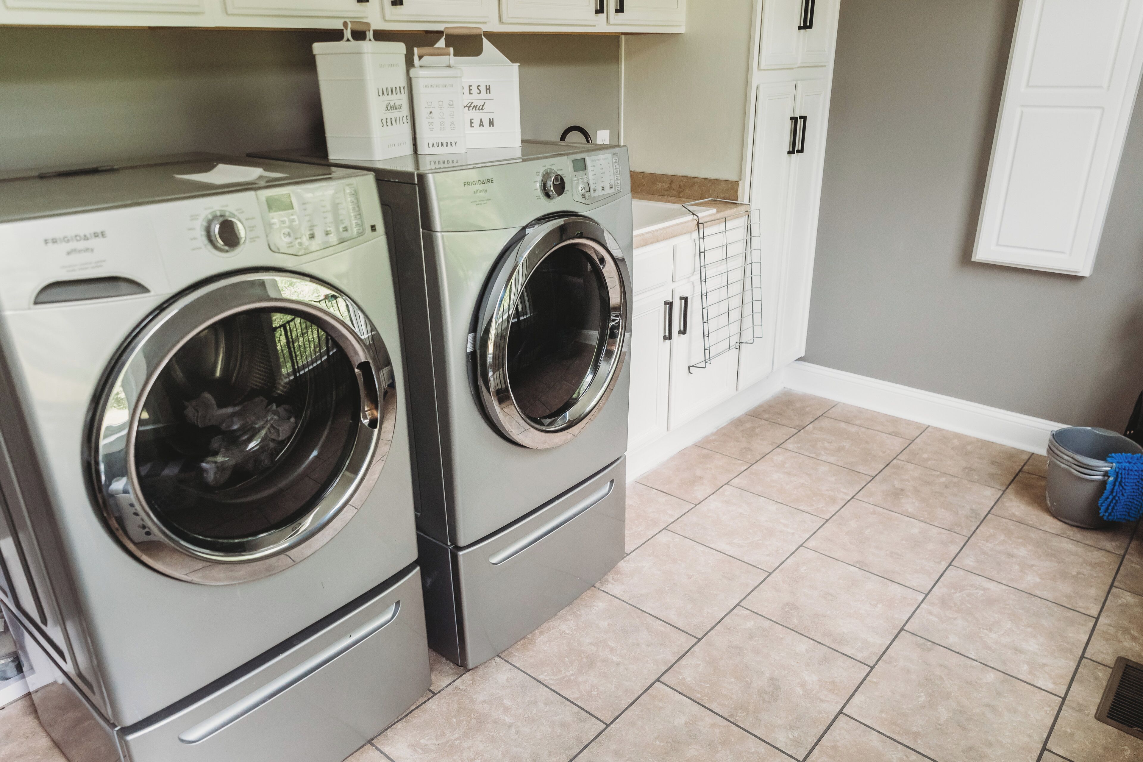 A laundry area with two front-loading washing machines, a drying rack, and a trash can.