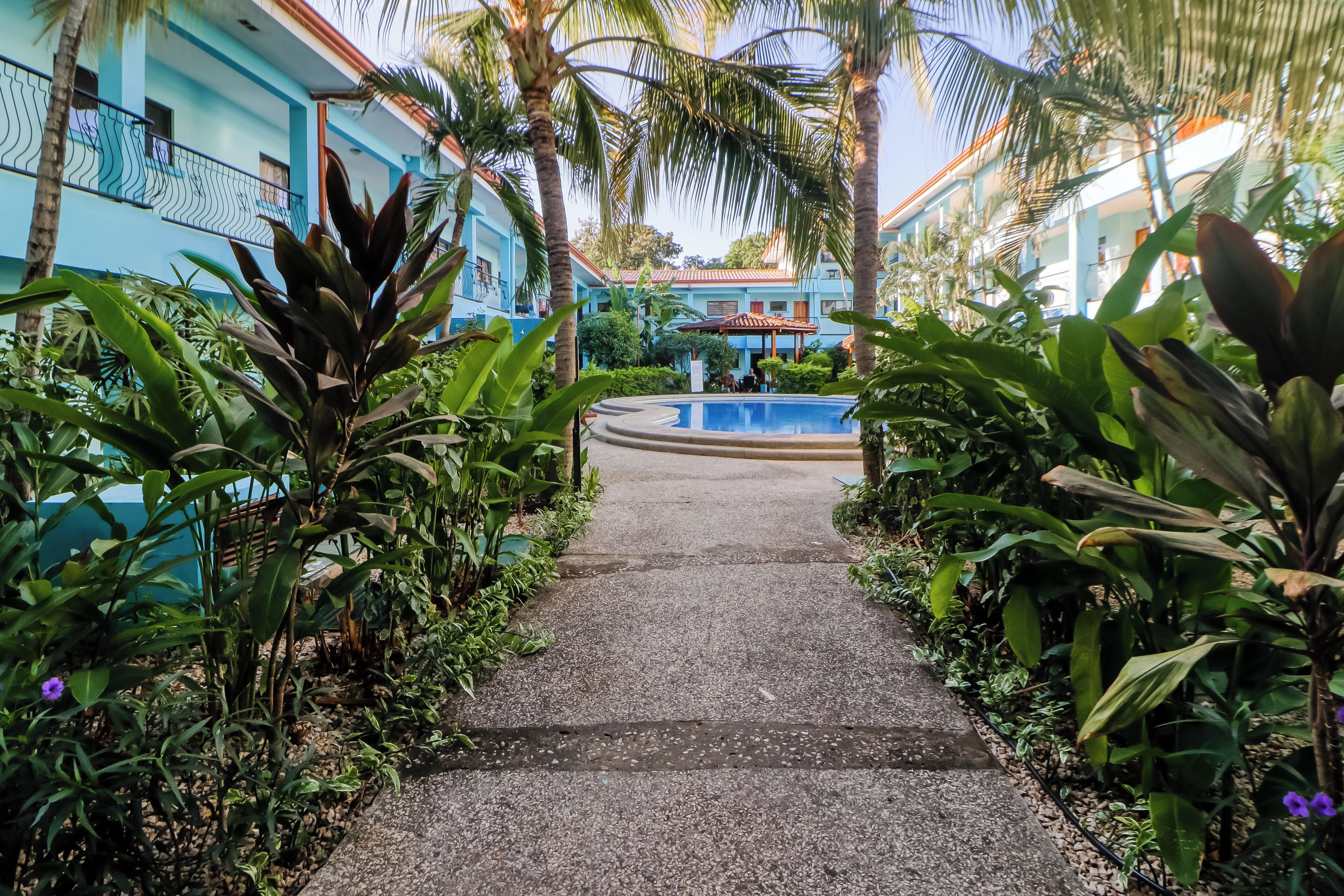 A pathway leading to a pool area surrounded by lush greenery and buildings with blue facades.