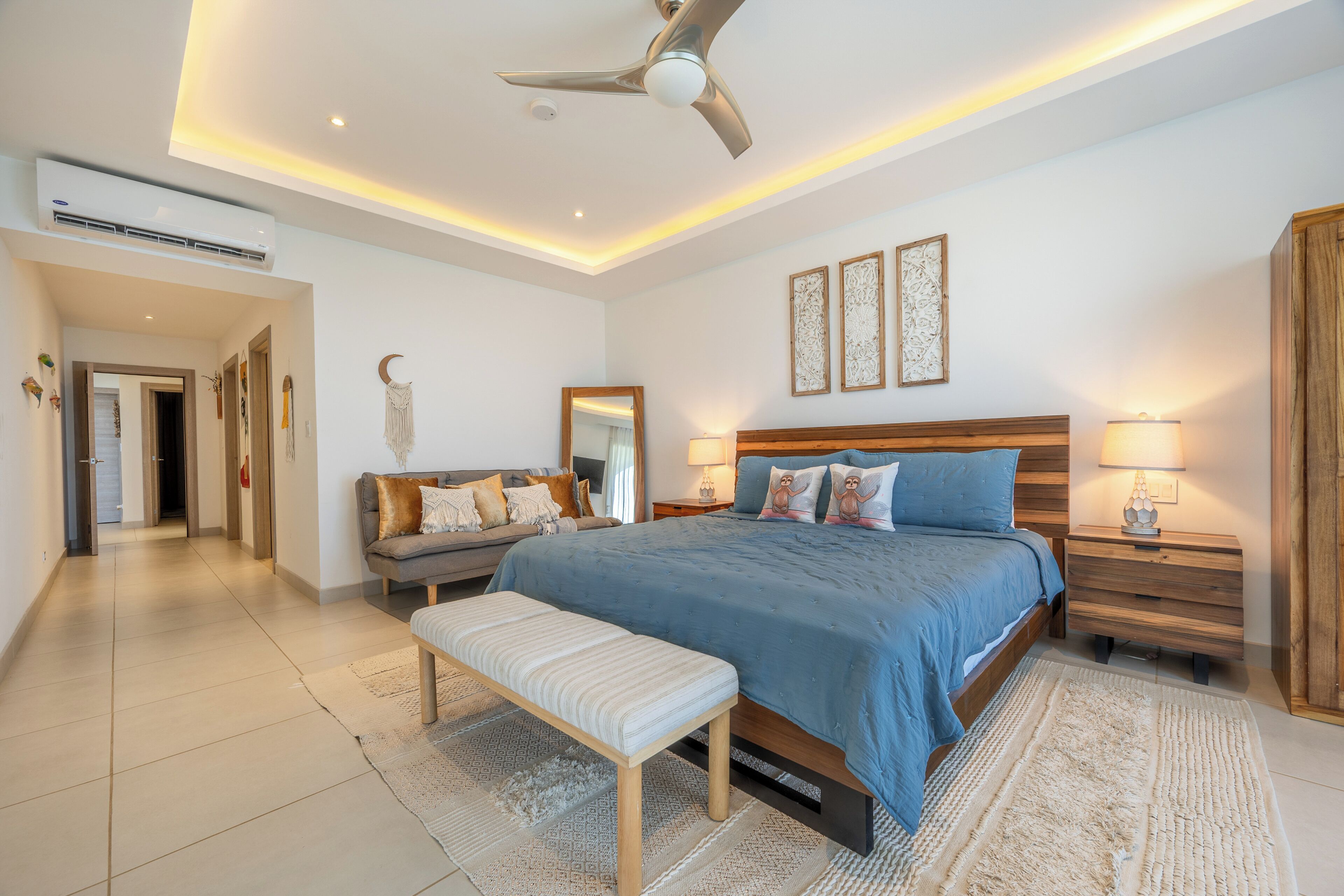 A modern bedroom with a large bed, a bench, a ceiling fan, and a nightstand with a lamp.