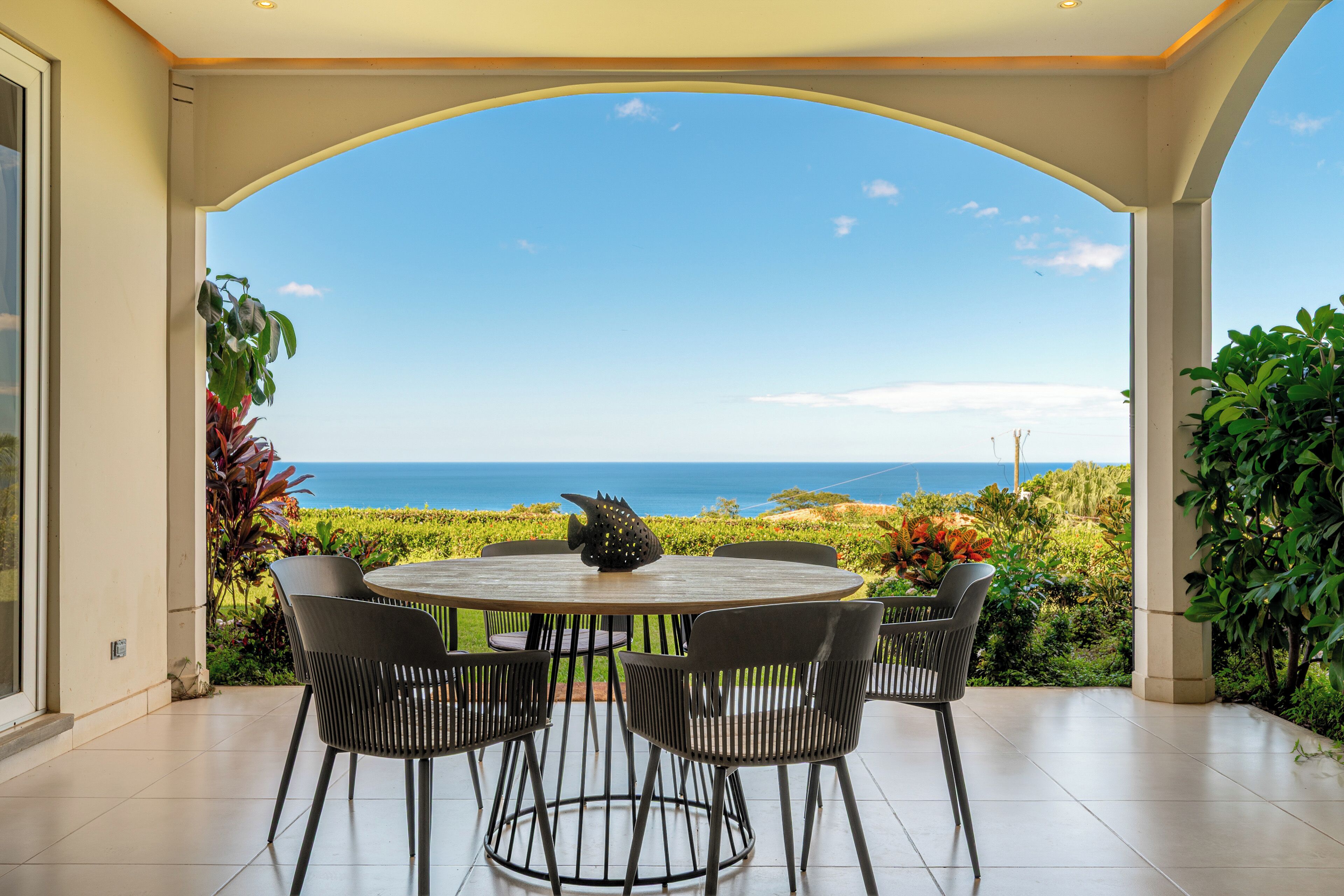 A covered patio with a round table and chairs, overlooking a scenic view of the ocean.