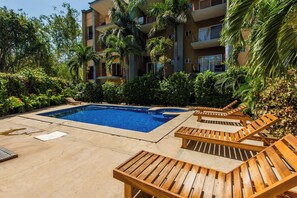 Une piscine entourée de chaises longues, bordée d’une végétation tropicale luxuriante et d’un immeuble à plusieurs étages doté de balcons.