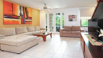 Amplia sala de estar con un sofá seccional color beige, una mesa de centro de madera y un televisor de pantalla plana sobre un mueble de madera. Hay dos ventanas que ofrecen luz natural y vistas a la vegetación exterior.