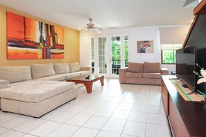 A spacious living room with a beige sectional sofa, a wooden coffee table, and a flat-screen TV on a wooden stand. There are two windows providing natural light and a view of greenery outside.