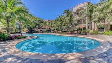 A swimming pool with a curved edge, surrounded by palm trees and lounge chairs, with a building in the background.