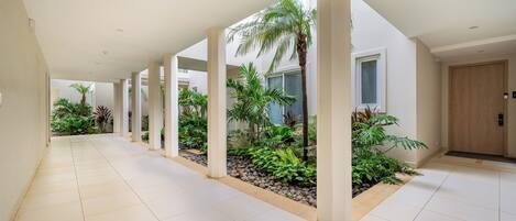 A corridor with tiled flooring, white walls, and columns supporting a roof. There are plants and a palm tree in the center.