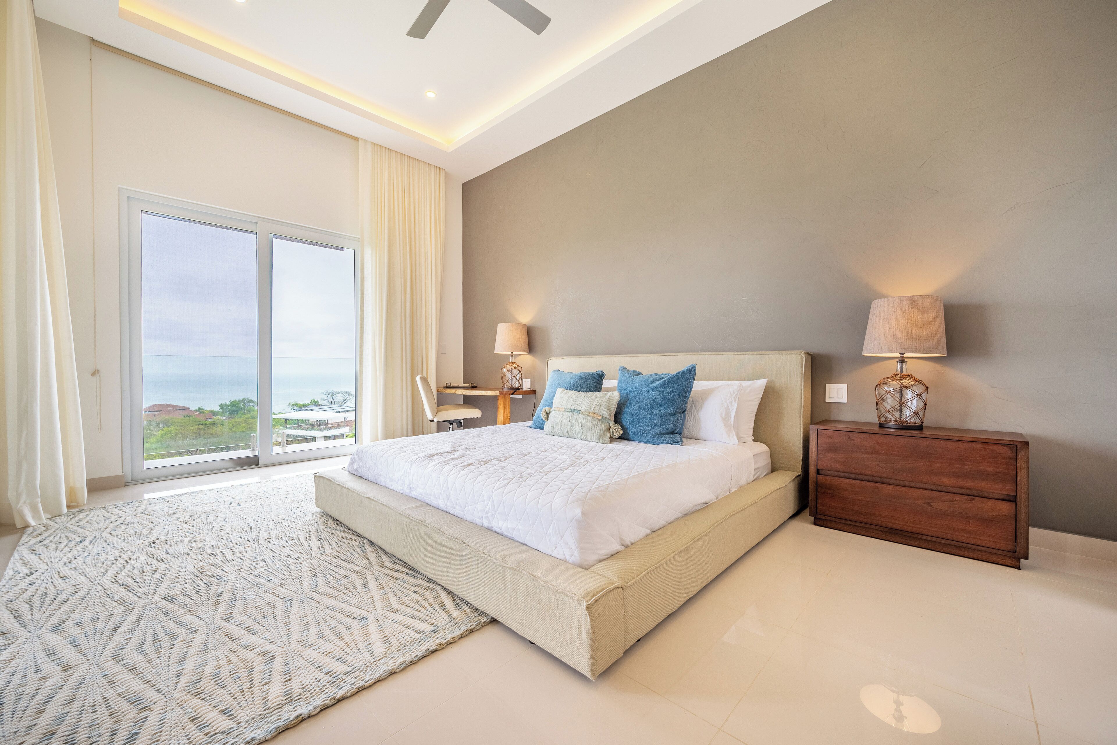 Quarto moderno com uma cama grande, duas luminárias de cabeceira, mesa de cabeceira de madeira e vista para o exterior por uma porta deslizante.