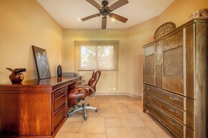 A room with a desk, chair, ceiling fan, window, and wooden cabinet.