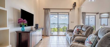 A spacious living room with a large sofa, a flat-screen TV, and a view of a balcony with a railing.