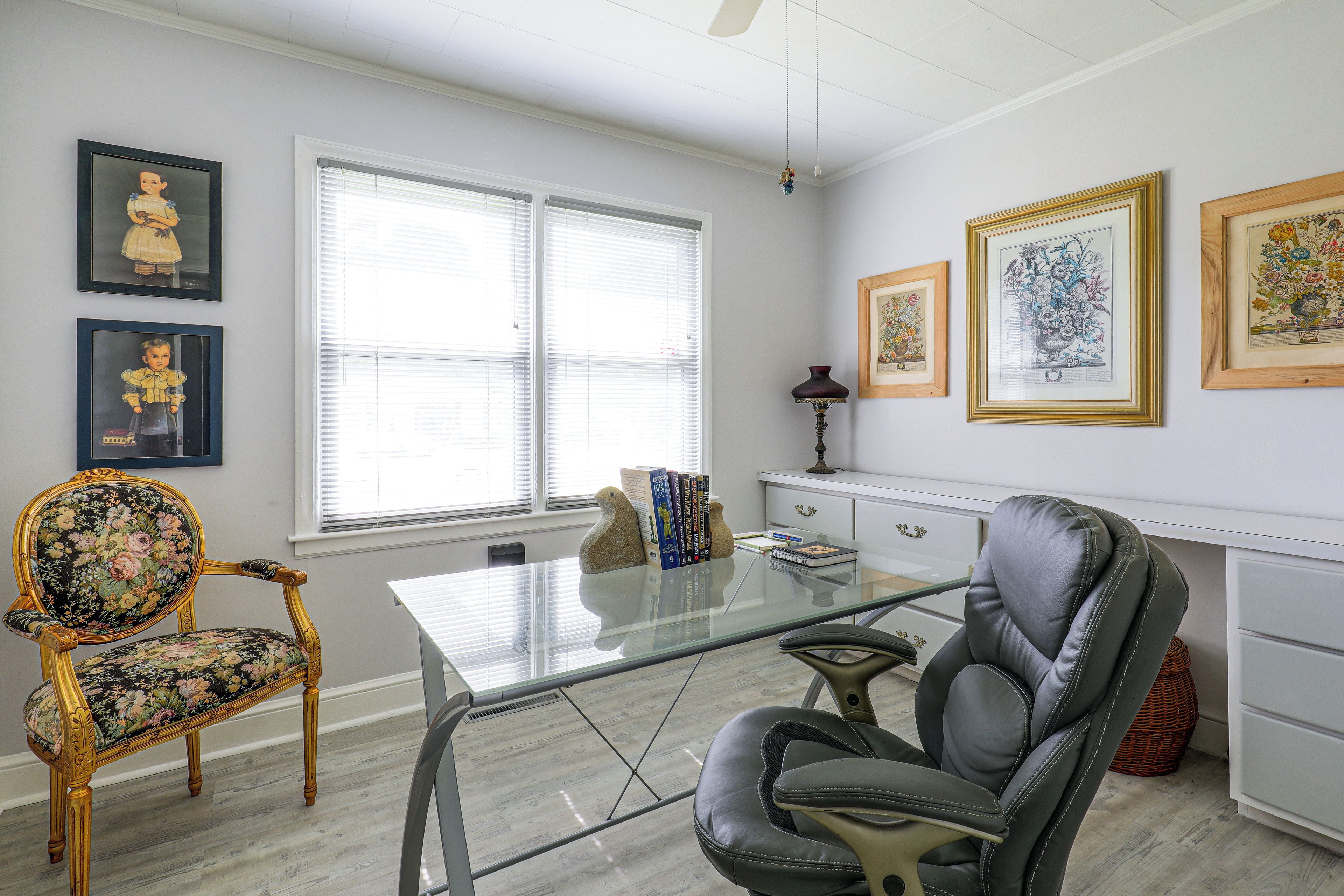 Una habitación con un escritorio de vidrio, dos sillas, un aparador blanco y cuadros en las paredes.