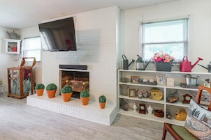 Una sala de estar con chimenea de ladrillo blanco, un televisor de pantalla plana montado arriba y una estantería llena de diversos objetos decorativos.