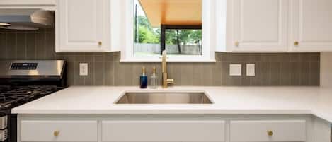 Una cocina con armarios blancos, un lavamanos de acero inoxidable y una ventana con vistas a la vegetación.