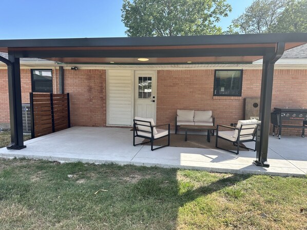 A covered patio area with a brick wall, a white door, and two chairs with cushions.