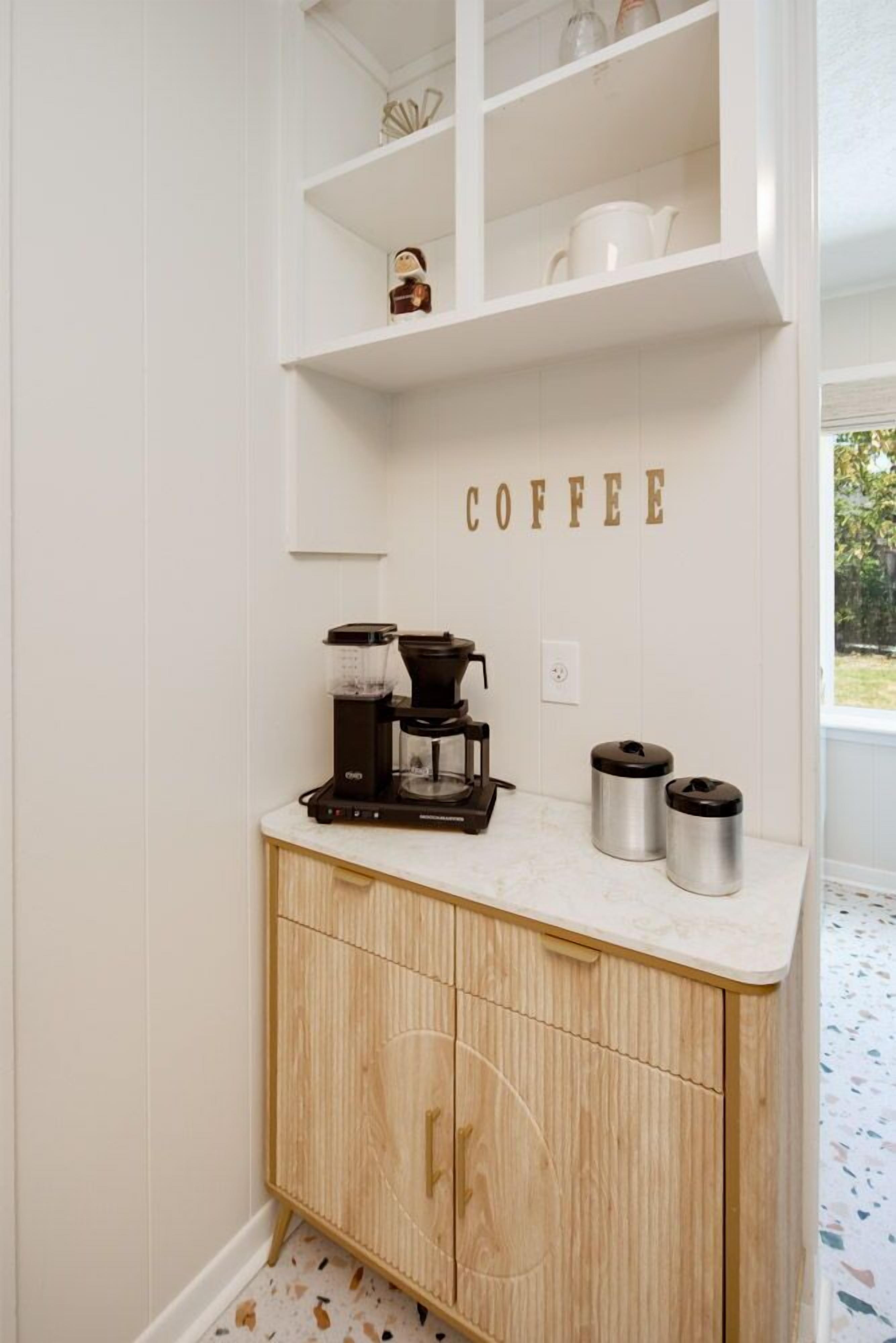 Eine Küche mit einer Kaffeestation, darunter eine Kaffeemaschine, zwei Thermoskannen und ein Regal mit Küchenutensilien.