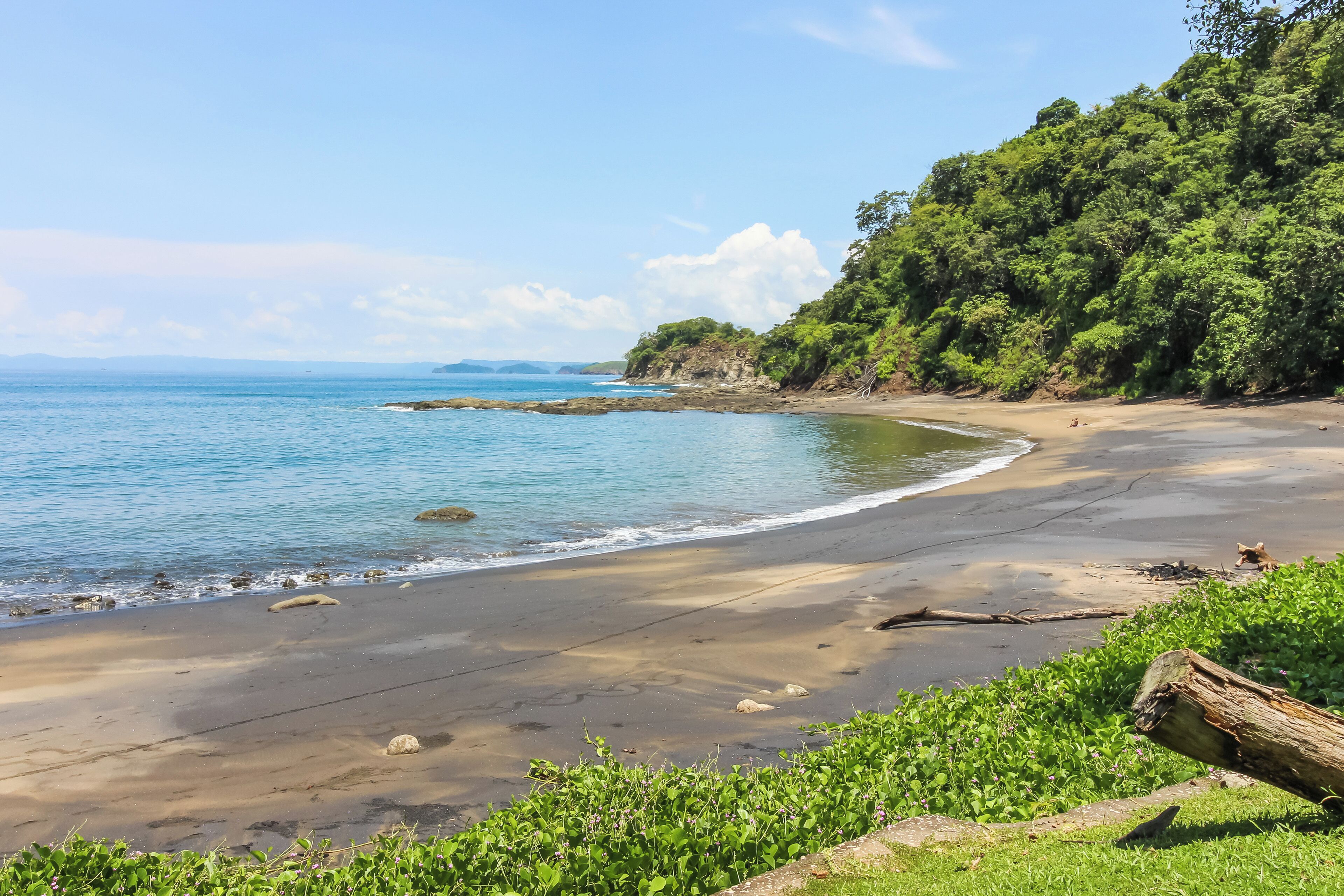 Una playa con costa rocosa, una extensión de arena y densa vegetación verde en una ladera.