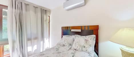 Quarto de hotel com cama, janela com cortinas, aparelho de ar condicionado e luminária.