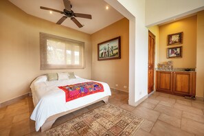 Un dormitorio con cama, ventilador de techo, ventana con persianas, un cuadro en la pared y una zona de cocina con armarios de madera.