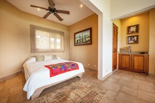 A bedroom with a bed, a ceiling fan, a window with blinds, a painting on the wall, and a kitchen area with wooden cabinets.