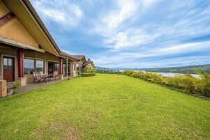 A house with a large lawn, a dining area with chairs and a table, and a view of a lake and hills in the distance.