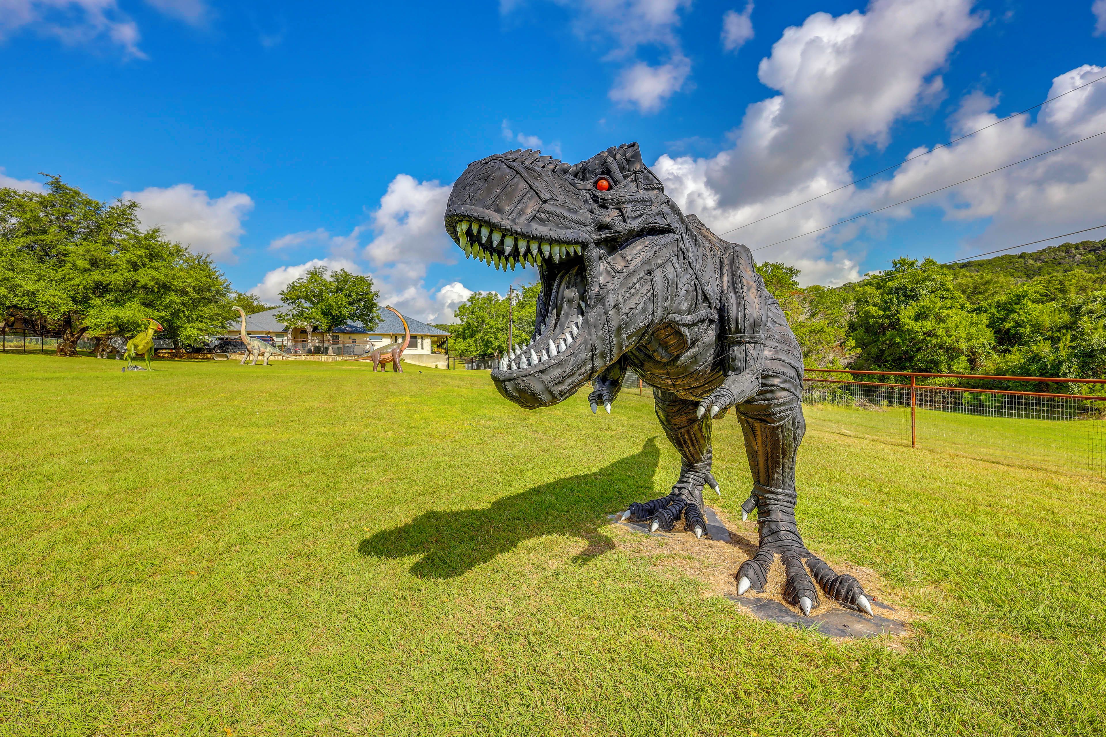 Una gran escultura de dinosaurio en un campo cubierto de hierba, con árboles y un cielo despejado al fondo.