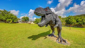 Una gran escultura de dinosaurio en un campo cubierto de hierba, con árboles y un cielo despejado al fondo.
