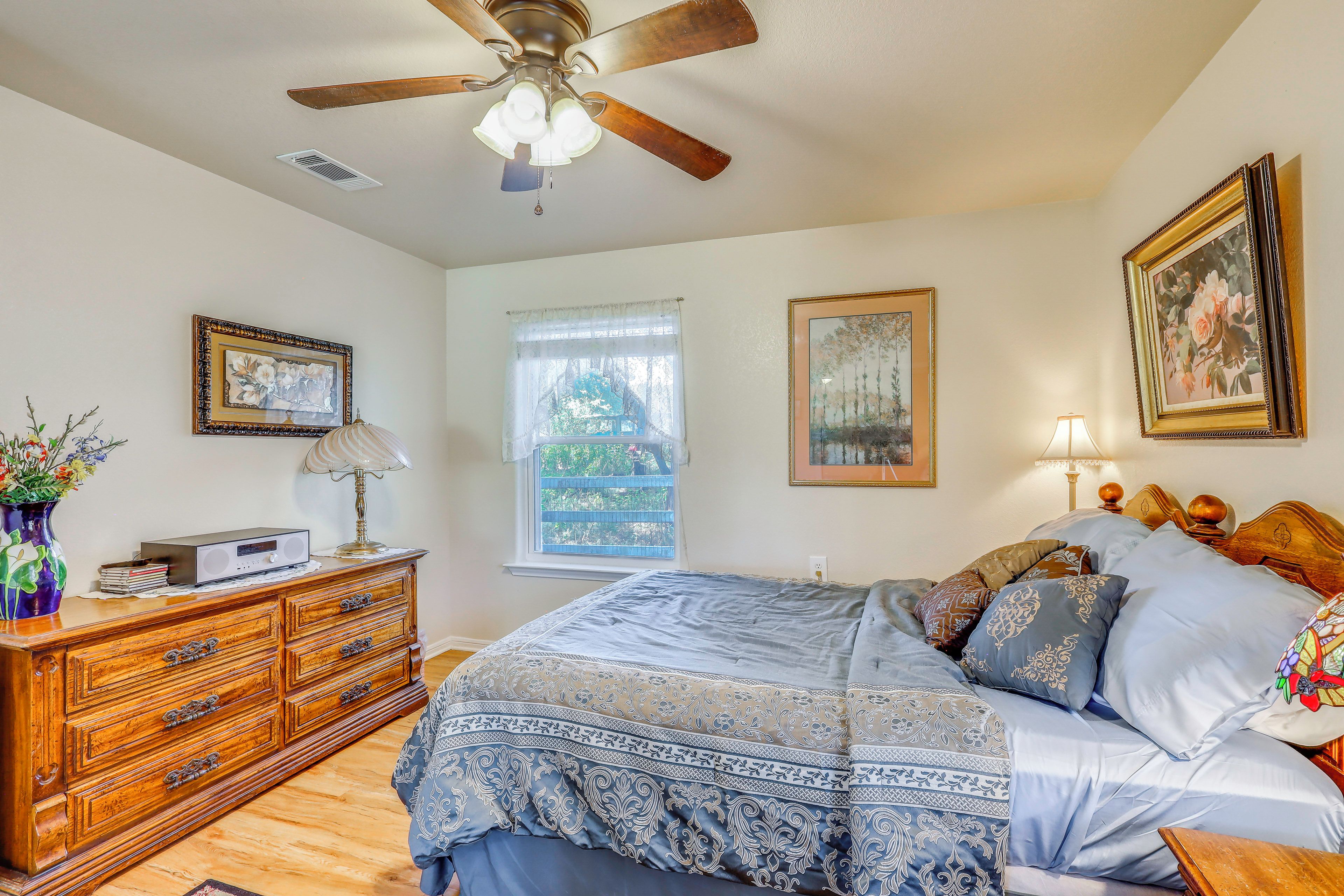 Une chambre à coucher comprenant un lit, une commode, un ventilateur de plafond, une fenêtre et des œuvres d’art.