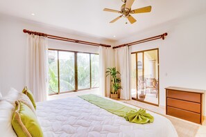 A bedroom with a large bed, a ceiling fan, a window with curtains, a plant, and a sliding door leading to a balcony. - Bright 3BR Sunrise Resort 2nd-floor Balcony (Tamarindo)