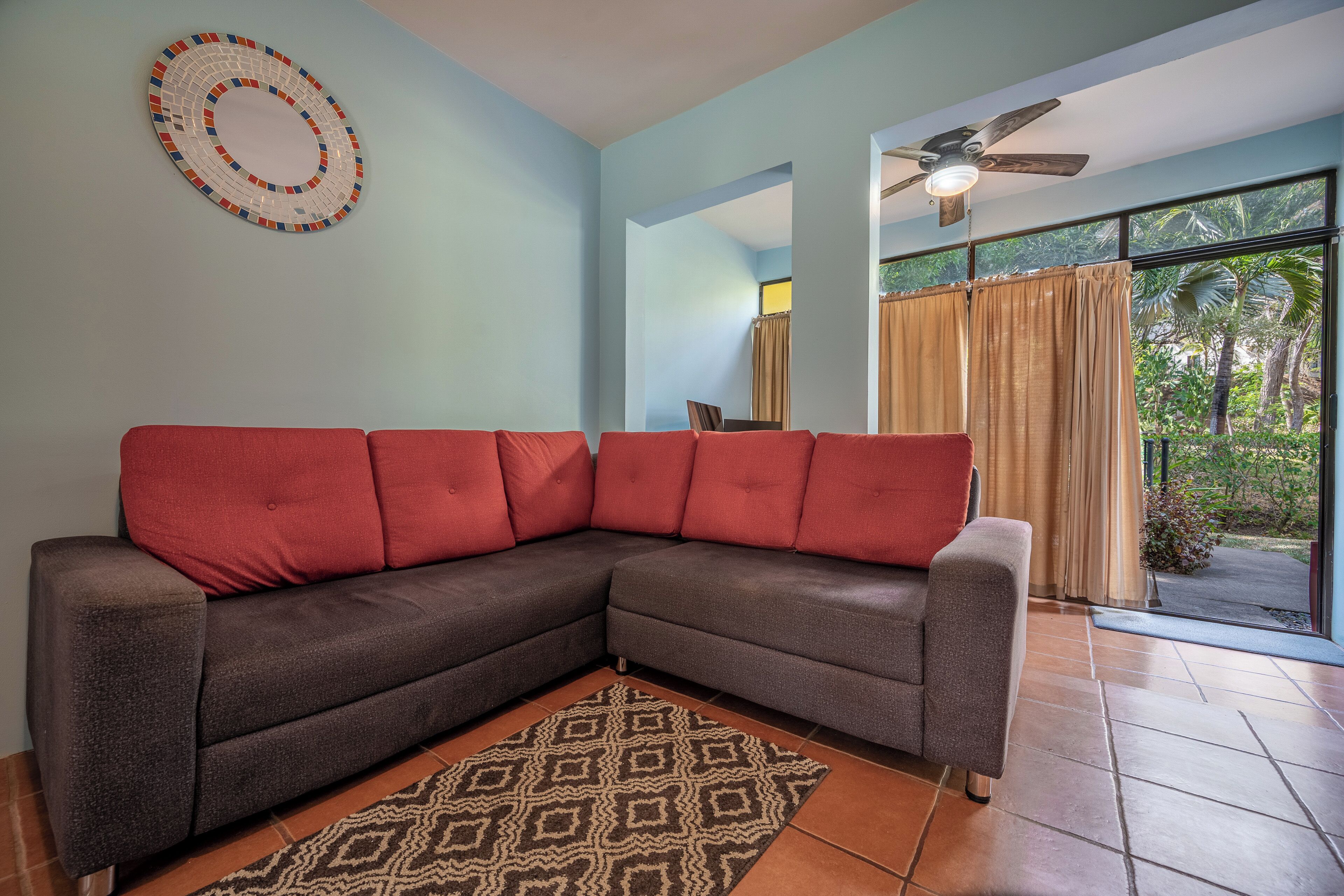 Una sala de estar con un sofá modular, un tapete estampado, un ventilador de techo y vistas a través de una ventana con cortinas.