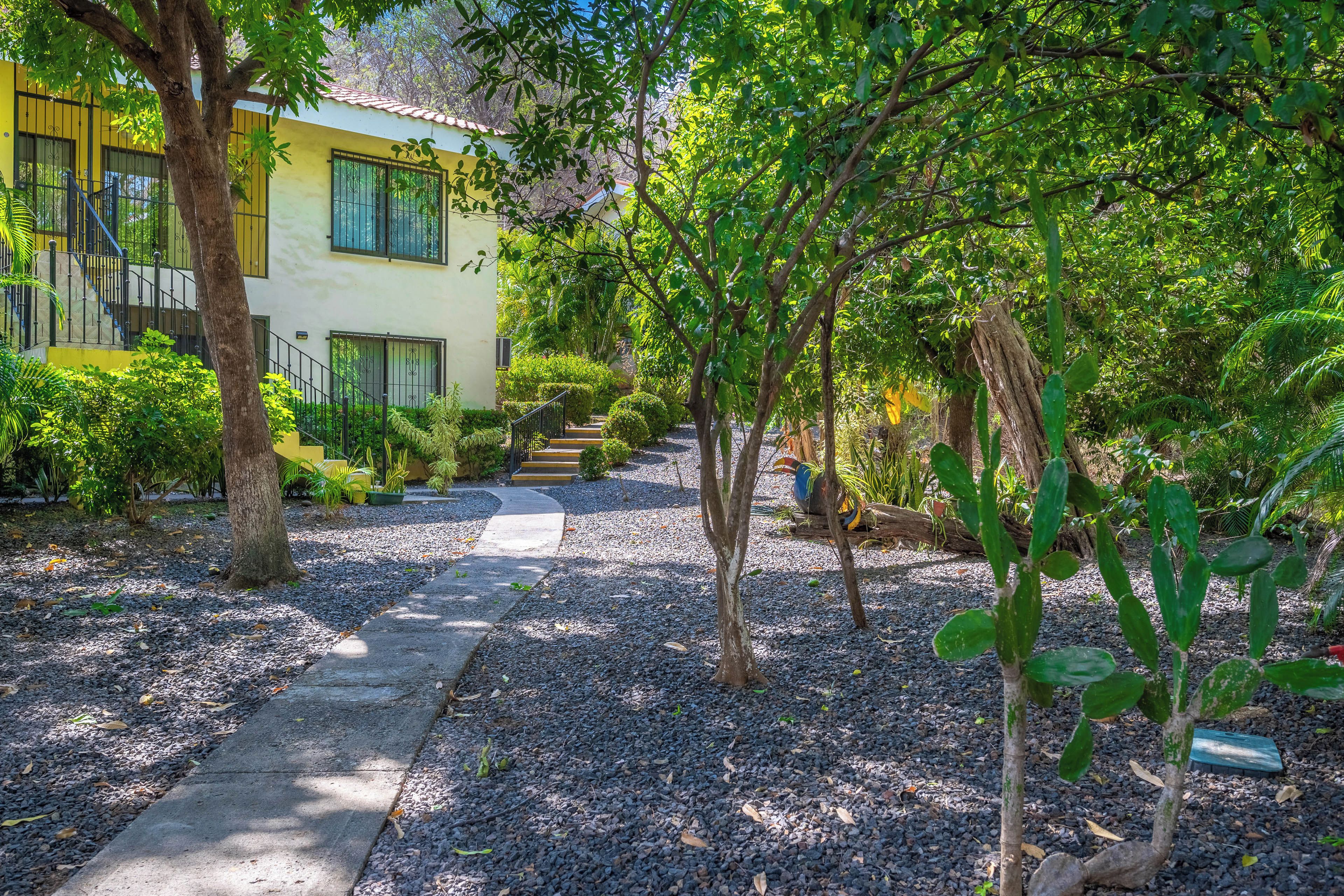 A pathway leading to a yellow building with a staircase, surrounded by greenery and trees.