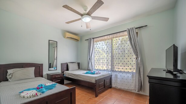 Una habitación con dos camas, un ventilador de techo, un equipo de aire acondicionado, un espejo, una ventana con cortinas y un televisor sobre un mueble.
