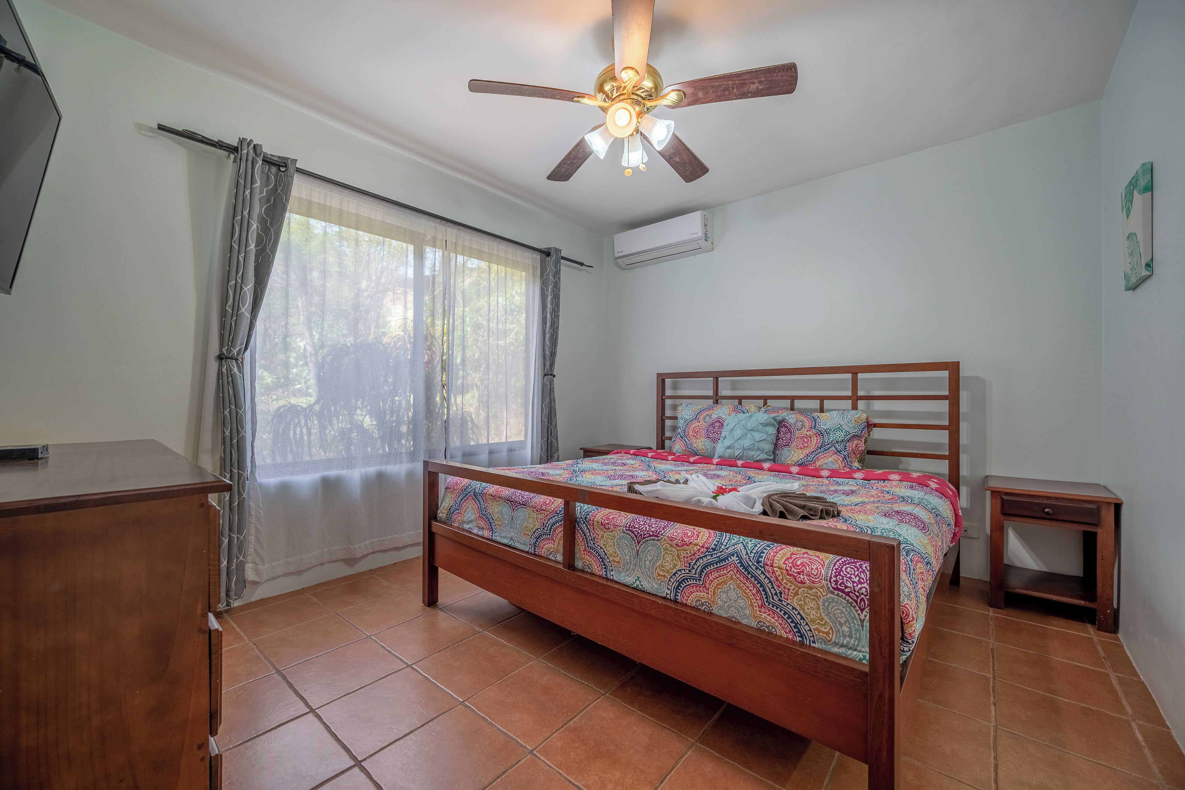 Une chambre avec un lit en bois, un ventilateur de plafond, une fenêtre avec des rideaux et un plancher carrelé.