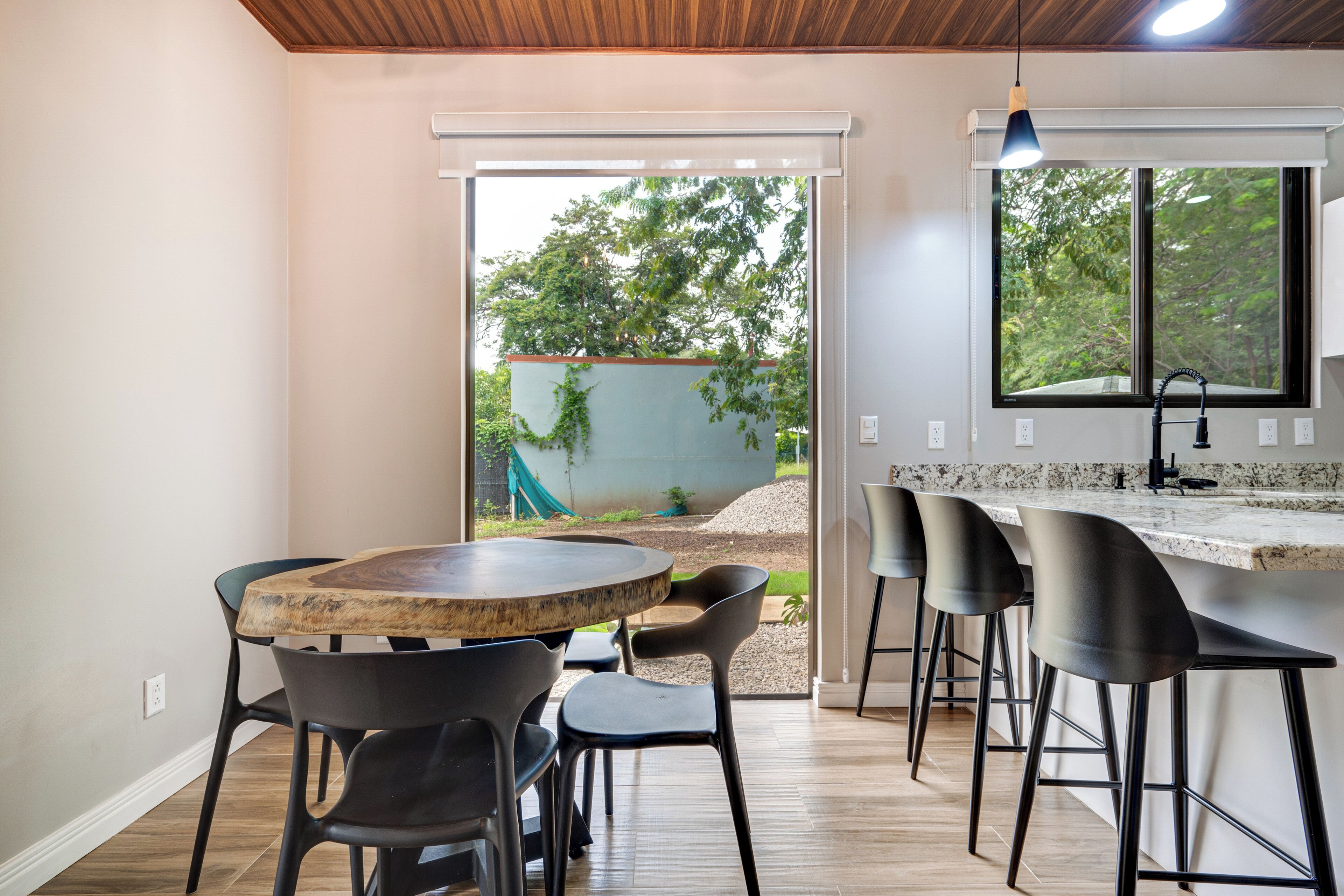 Une cuisine moderne avec une table en bois et des tabourets de bar, une grande fenêtre donnant sur un paysage verdoyant et un plafond avec des poutres en bois.