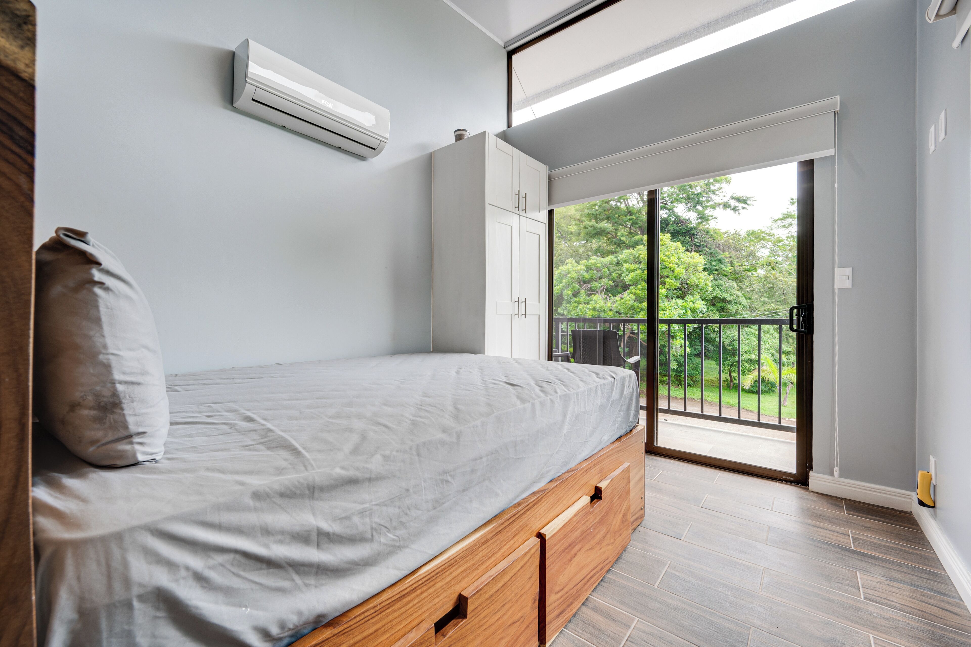 A bedroom with a bed, an air conditioner, a sliding door to a balcony, and a view of greenery.