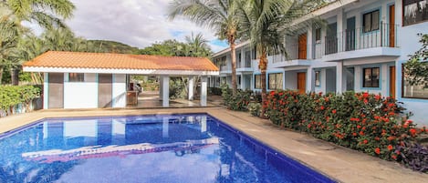 Un hotel con piscina, zona de estar techada al aire libre y jardín con flores rojas.